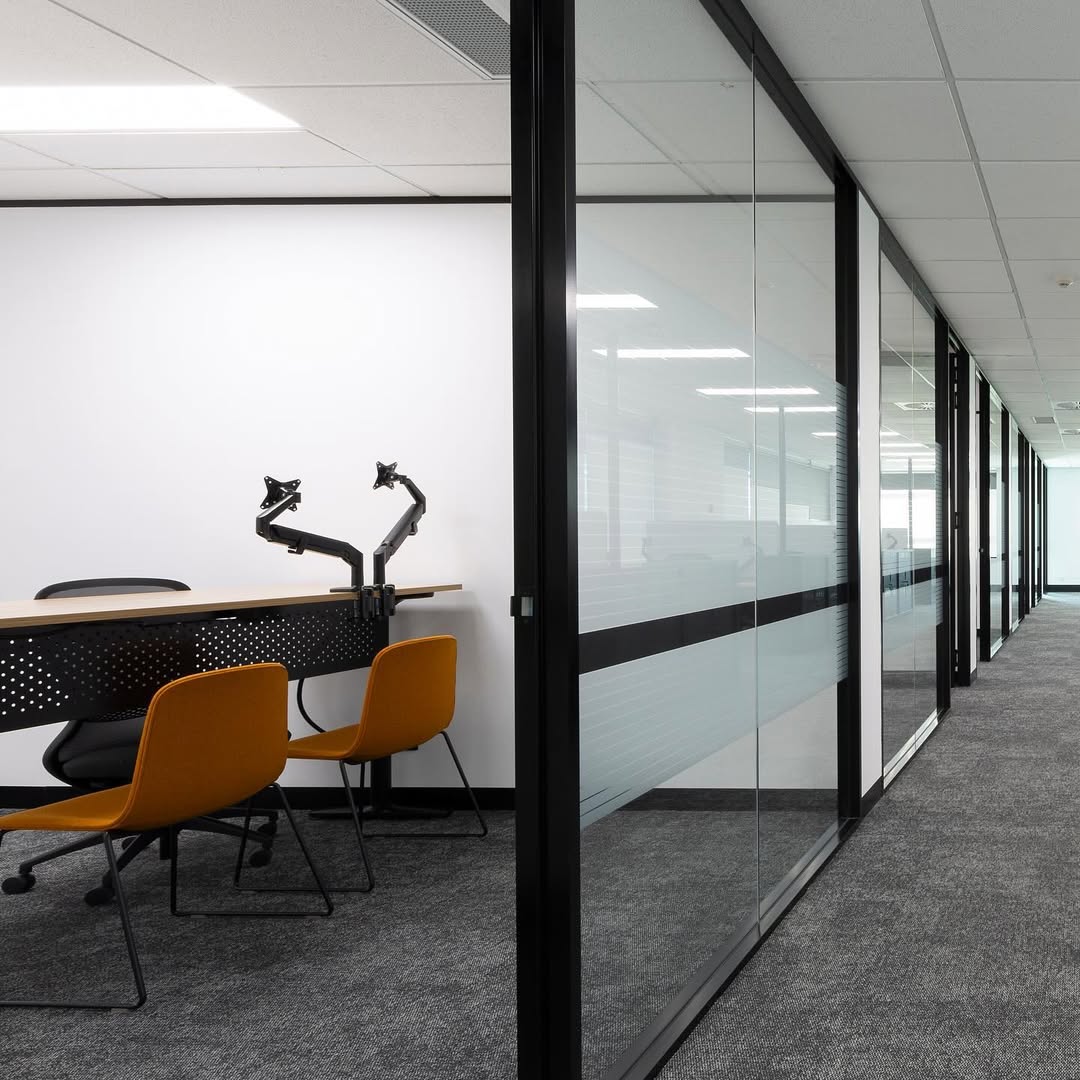 Modern office with gray carpet, long corridor, glass walls, and a meeting area featuring a wooden desk, black monitors, and orange chairs.