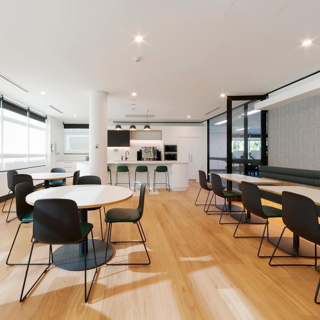 Spacious, modern and clean office kitchen with wood flooring, round tables, and black chairs. A white island counter with stools and industrial lighting enhances the sleek design.