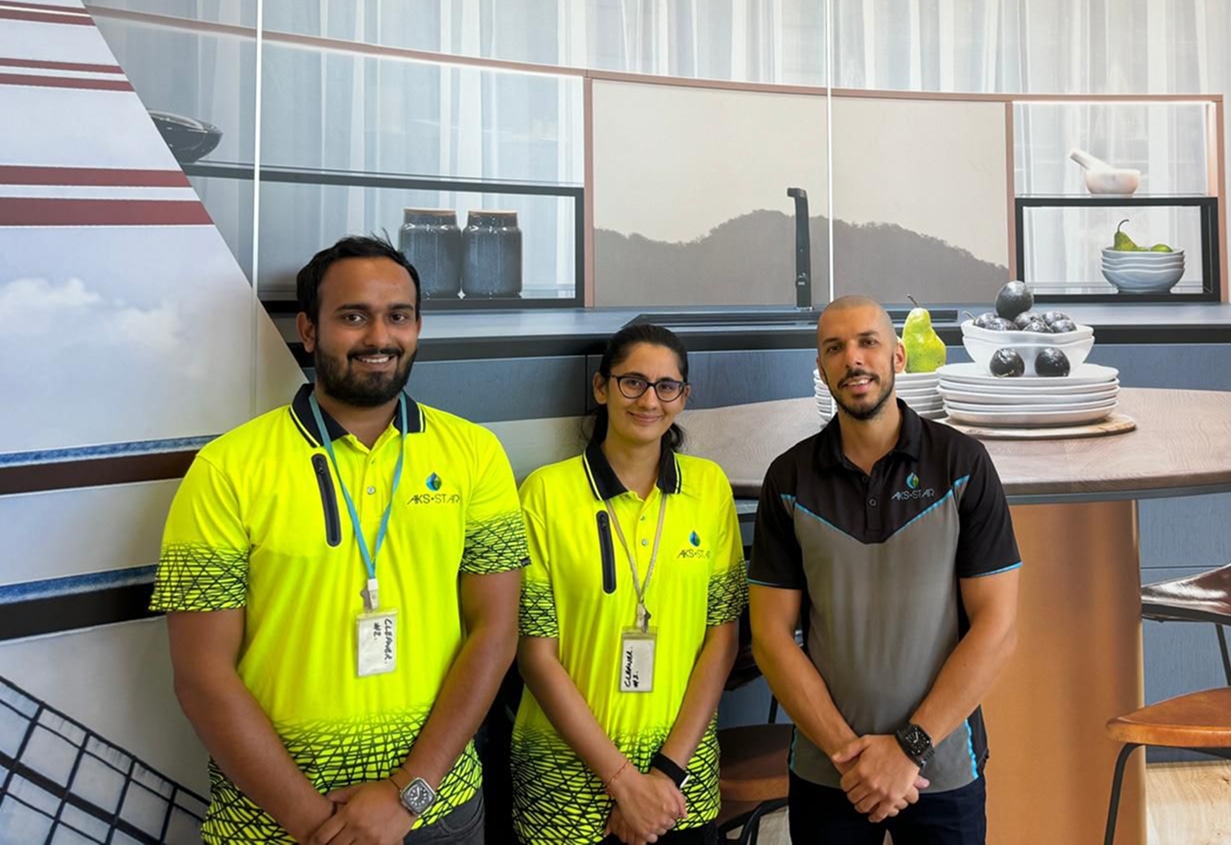 Three people smiling in cleaning uniforms stand indoors in front of a wall with a kitchen backdrop. The mood is friendly and professional.